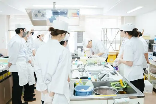 白衣姿の調理師たちが調理実習室で指導を受けながら作業している様子